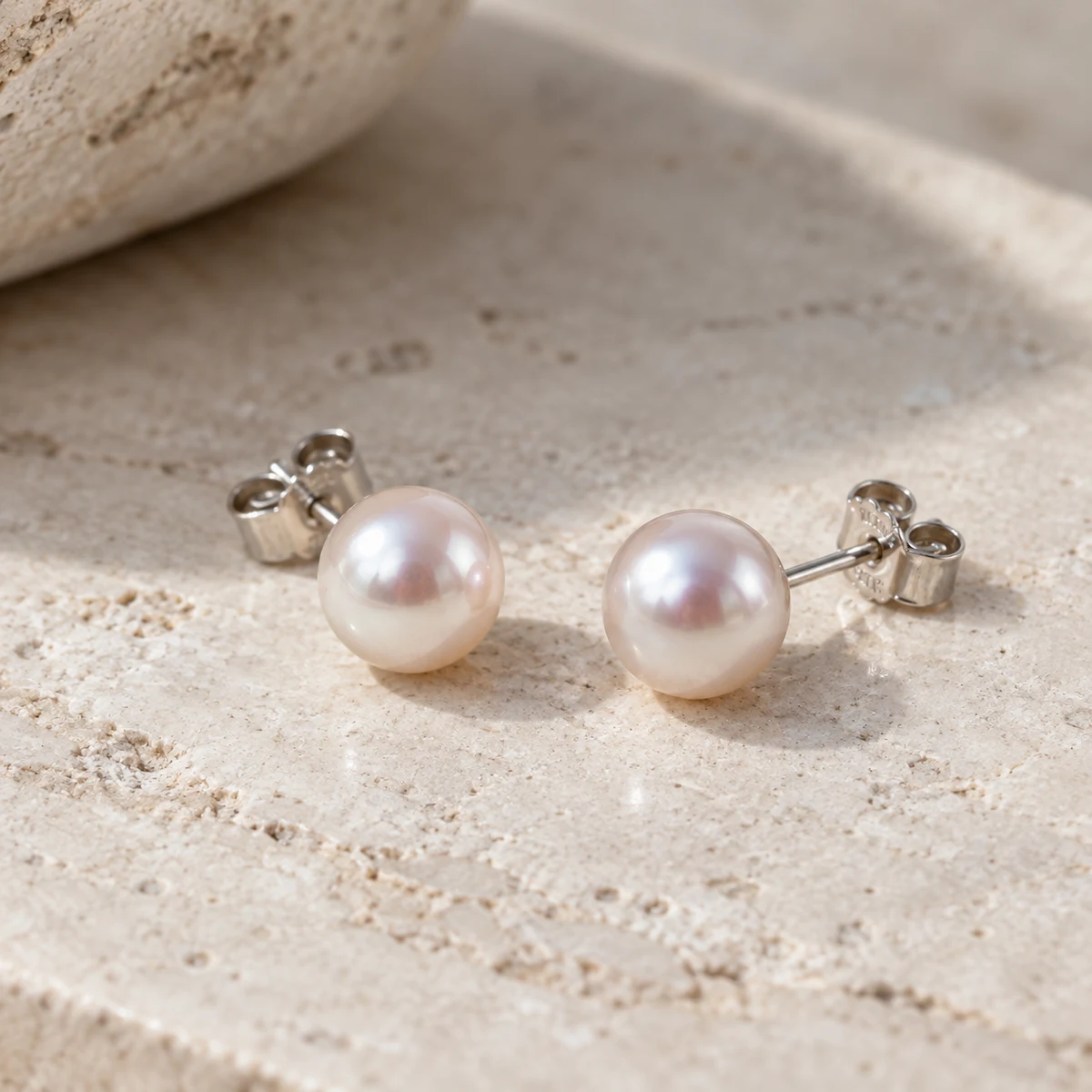 Matched pair of white Akoya-style pearl stud earrings on pale limestone; concept photograph