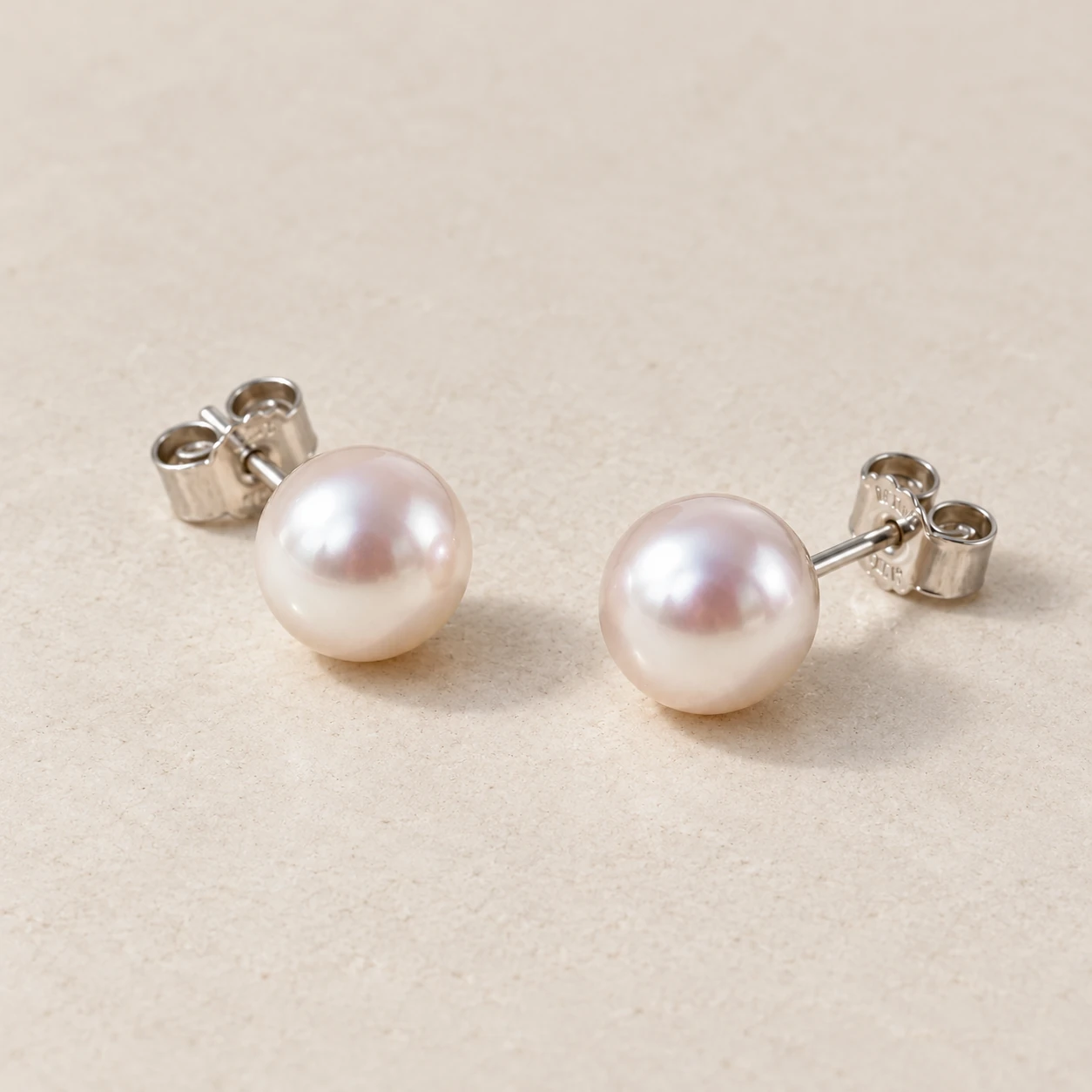 Pair of white Akoya-style pearl stud earrings on pale warm-grey stone; concept photograph