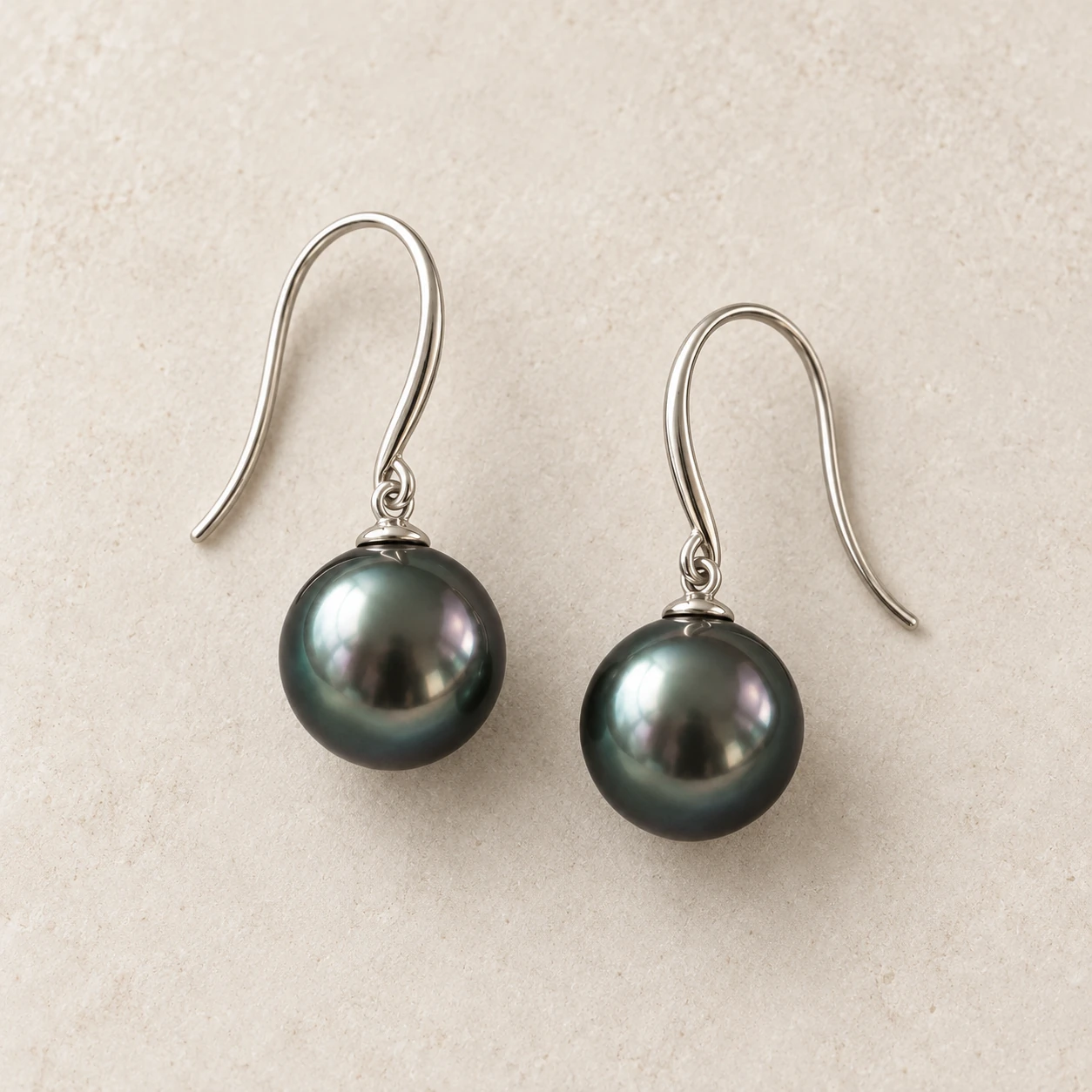 Pair of dark Tahitian-style pearl drop earrings on pale warm-grey stone; concept photograph